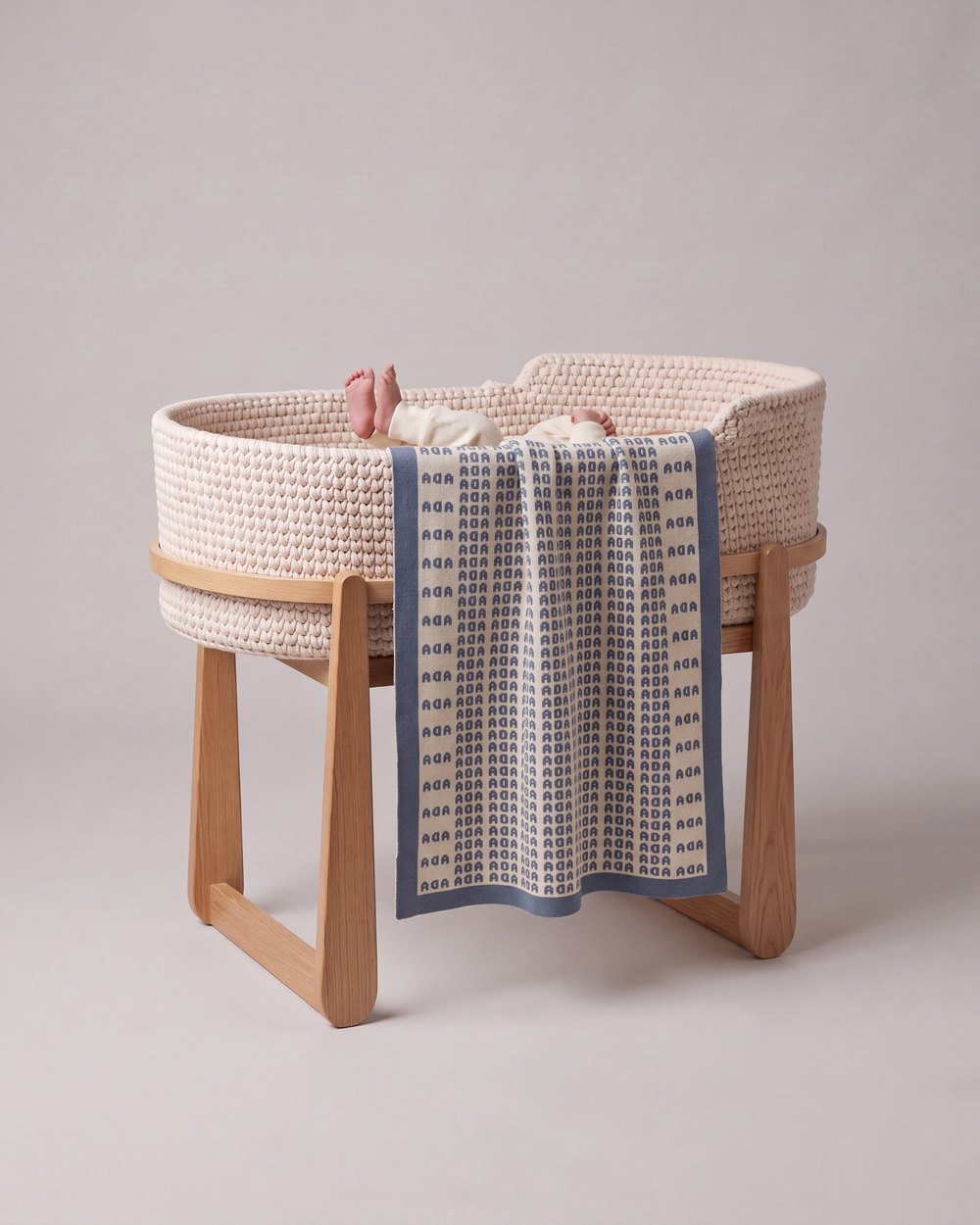 A Heir blanket with the name Ada knitted in blue, draped over a woven moses basket on an oak stand