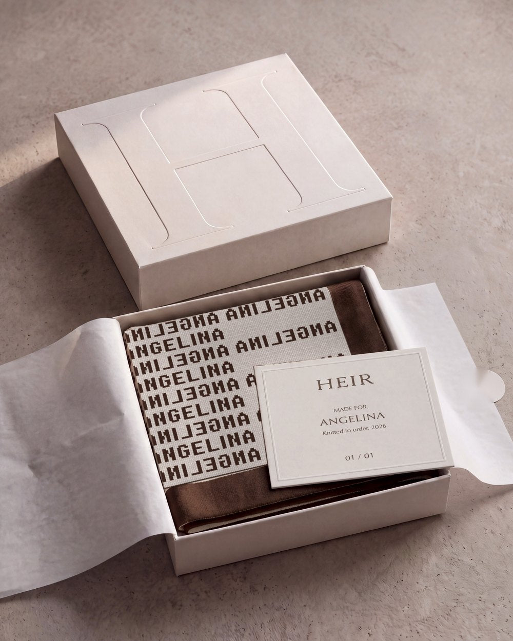 The open Heir box: an Angelina blanket in chocolate inside tissue, with a card reading made for Angelina, beside the embossed lid