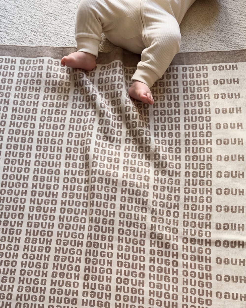 A baby's bare feet on a Heir blanket with the name Hugo knitted in taupe, laid on a carpet