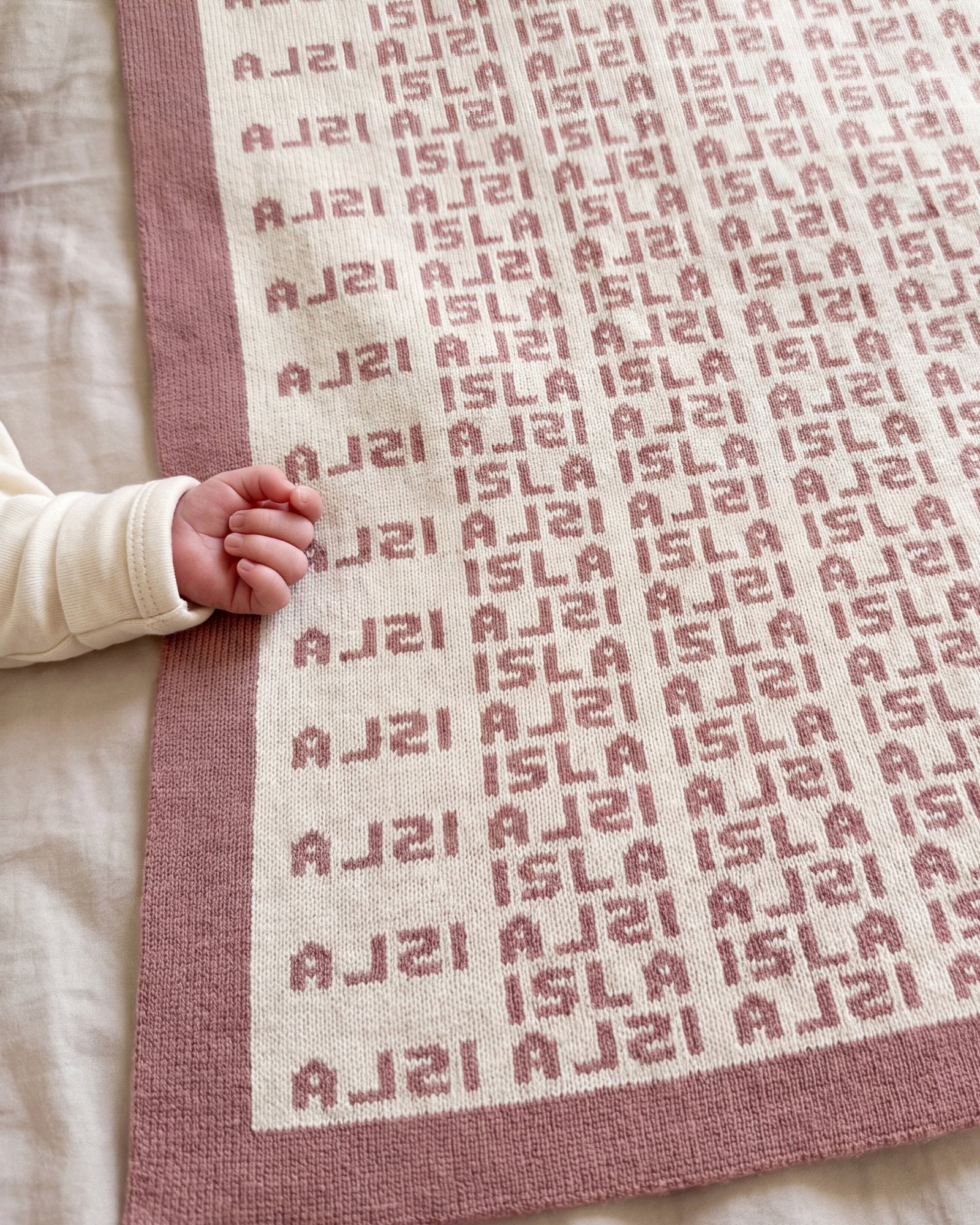 A newborn's hand at the rose border of a Heir blanket with the name Isla knitted in rose