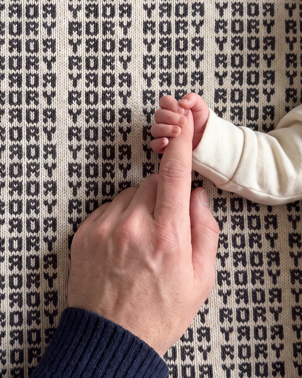 A baby's hand gripping a father's finger on a Heir blanket with the name Rory knitted in charcoal
