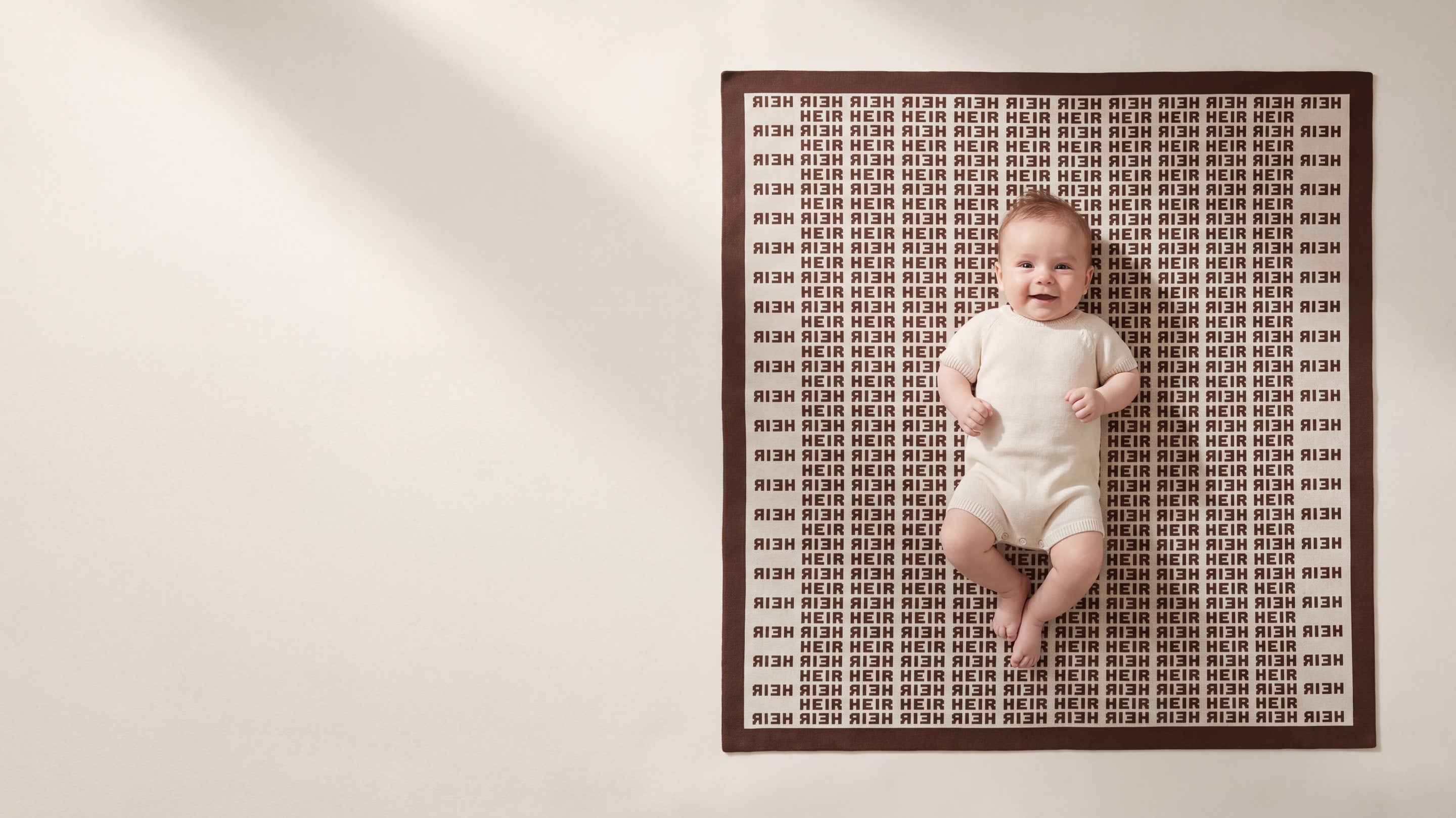 A smiling baby lying on a Heir blanket with the name knitted in rows of chocolate brown on cream